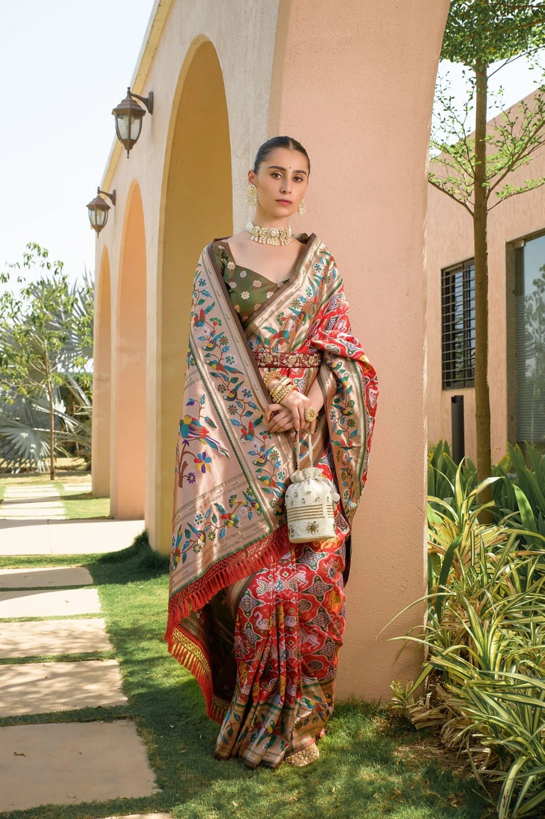 Woman in a colorful saree standing outdoors with a building and greenery in the background